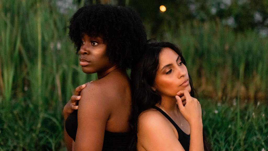 Two women standing back-to-back in a natural setting with greenery.